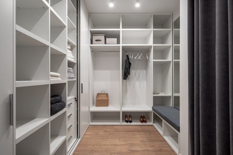 Trendy white dressing room with luminous lamps and a parquet on the floor. There are lockers, shelves and hangers with clothing, shoes, tall mirror, soft seat with niches, gray curtains. Horizontal.