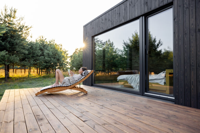 Young woman resting on sunbed and reading on a tablet on the wooden terrace near the modern house with panoramic windows near pine forest. Concept of solitude and recreation on nature